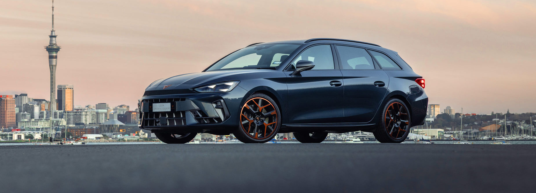 A dark grey CUPRA wagon with bronze wheels parked near the Auckland waterfront